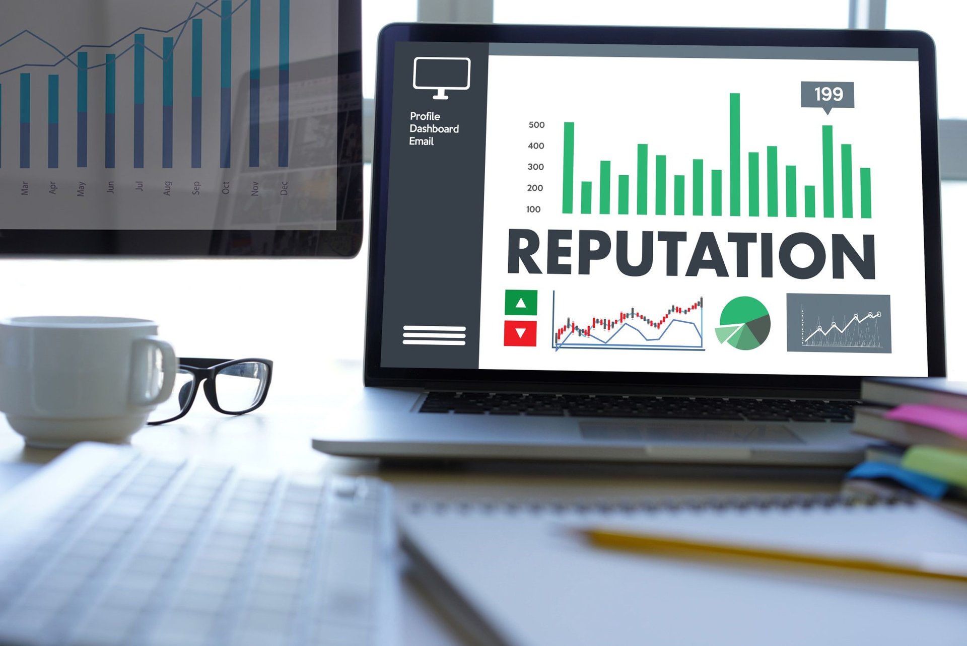 A laptop showcasing graphs labeled "REPUTATION" sits alongside a monitor displaying a bar graph, surrounded by essential tools like glasses and notebooks on the desk, perfect for online reputation management.