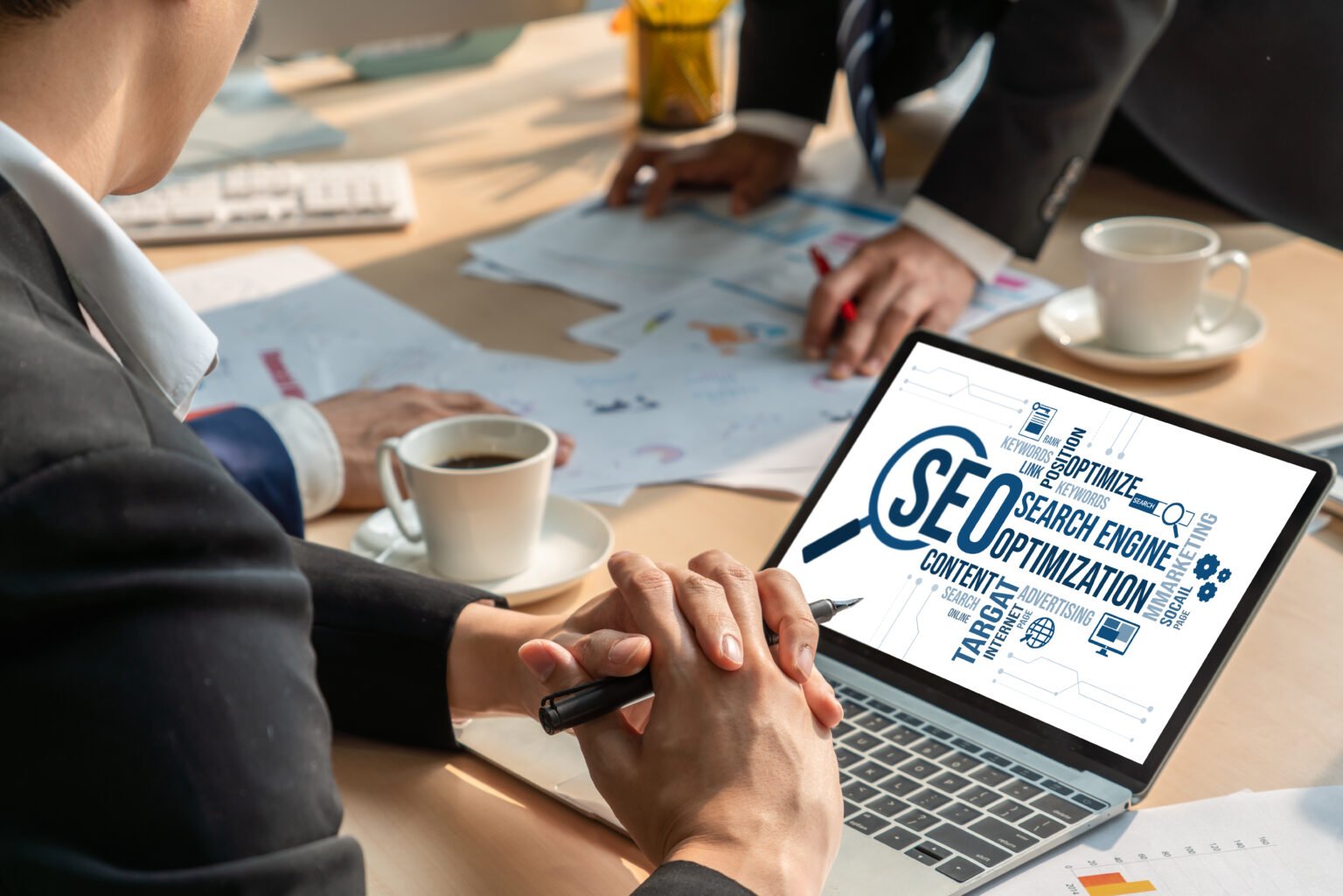A person in business attire attentively views an SEO presentation on their laptop, surrounded by colleagues, coffee cups, and scattered papers. This scene prompts the question of why investing in such strategies is worth it, despite being potentially expensive.