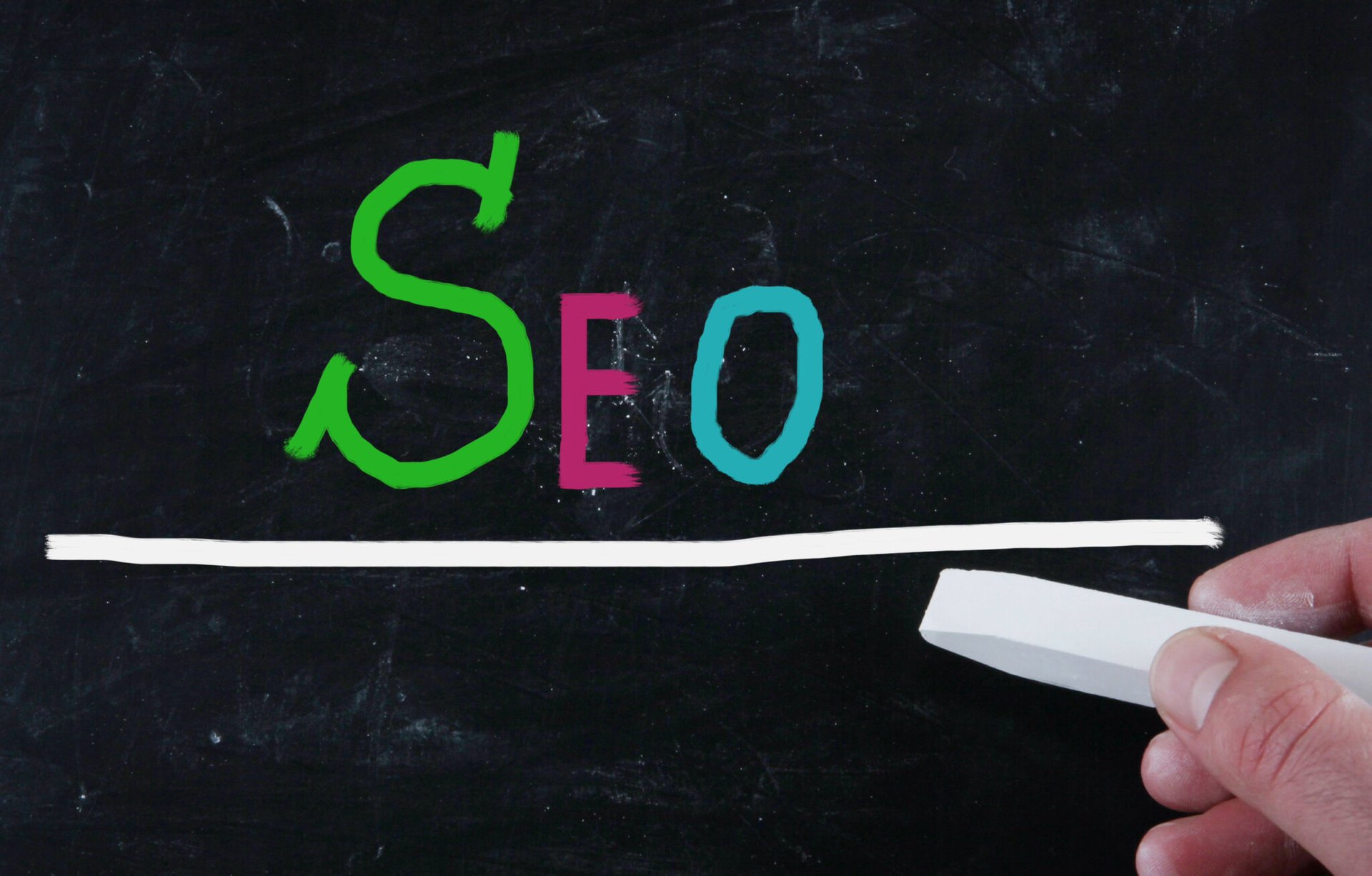 A hand holding a white chalk piece draws a line under colorful "SEO" letters on a blackboard, illustrating the foundation of effective SEO services.