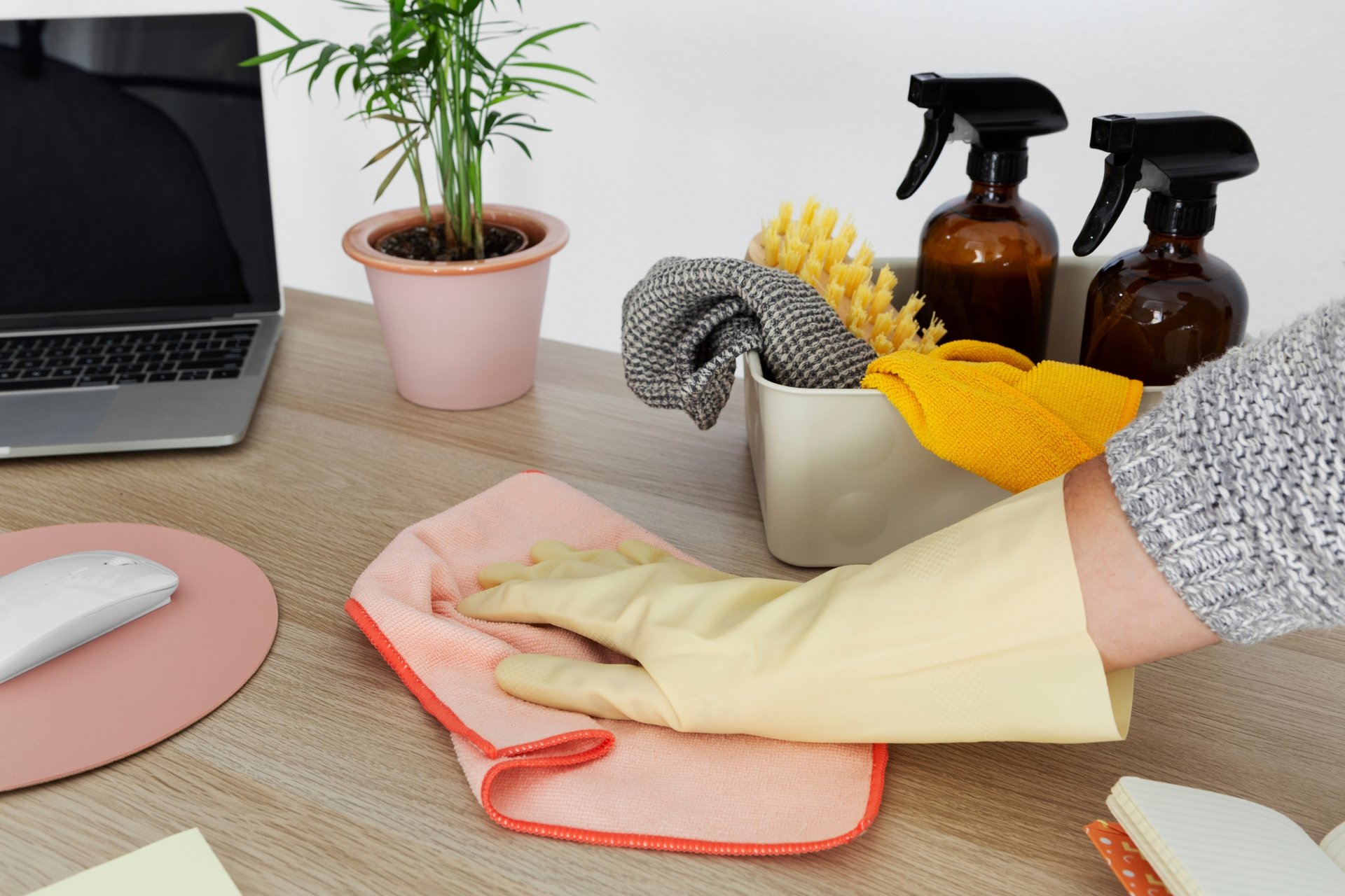 The Real Cost of DIY Home Cleaning vs. a Professional Service 1 Yellow-gloved hand wiping wooden desk surface with pink cloth beside laptop and cleaning caddy