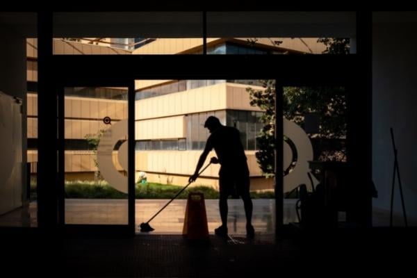 Shopping Centres 4 AustClean shopping centre after hours cleaning – silhouette of cleaner mopping floor near large glass doors with caution sign in view, modern buildings outside; professional retail and commercial cleaning for safety and presentation in Australia.