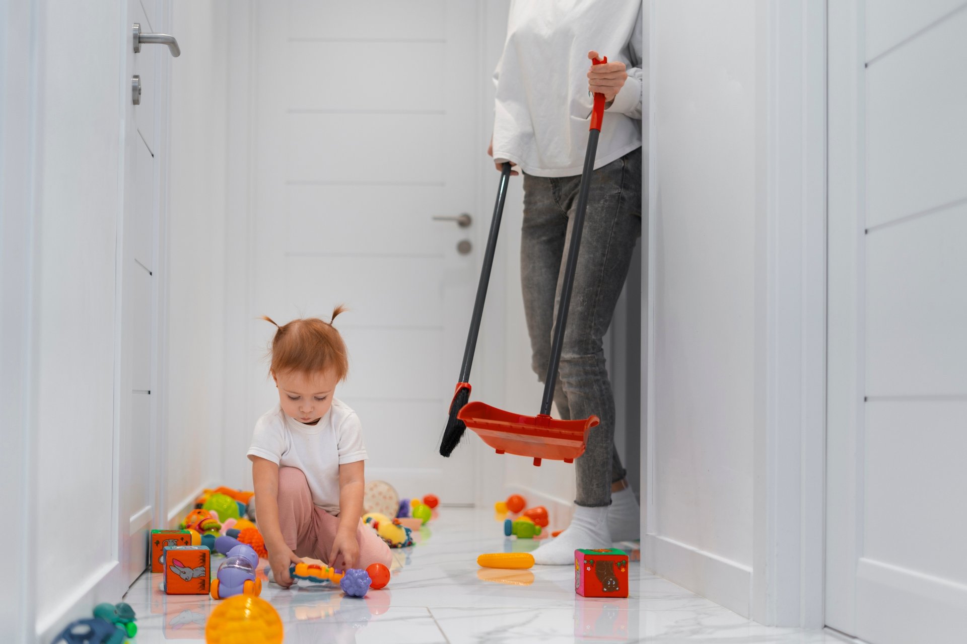The Busy Parent's Guide to a Clean Home in Mountain Creek 1 Toddler playing with colourful toys on hallway floor while adult holds broom ready for child-safe house cleaning Mountain Creek