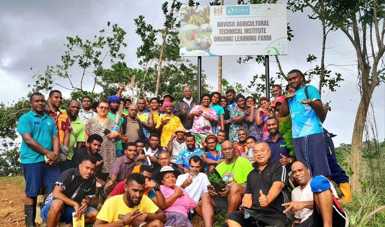 A large group of people pose together outdoors in front of a sign that reads "Navuso Agricultural Technical Institute Organic Learning Farm.