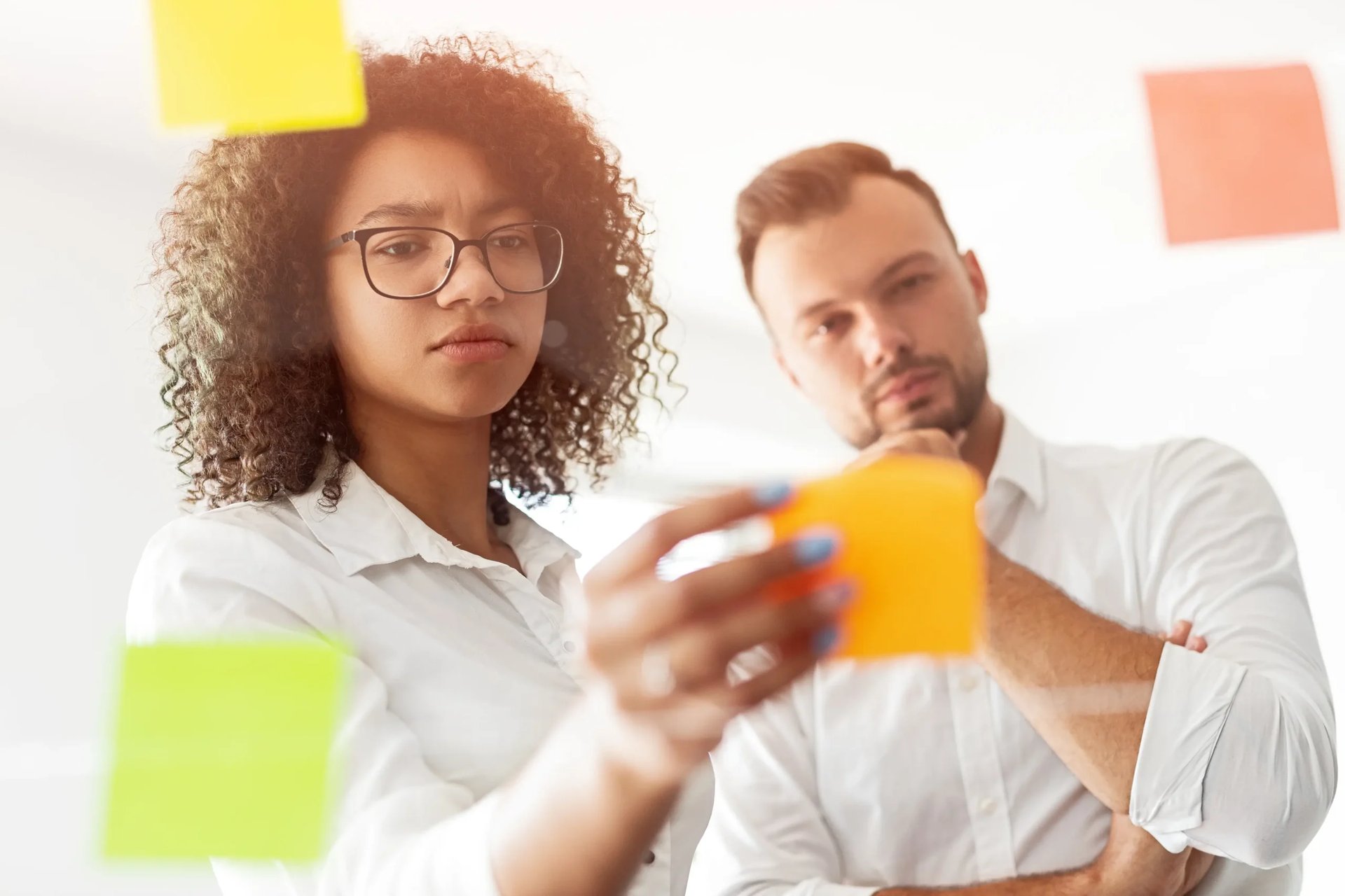 Black woman attaching sticky note on glass while pondering over creative strategy during meeting in office