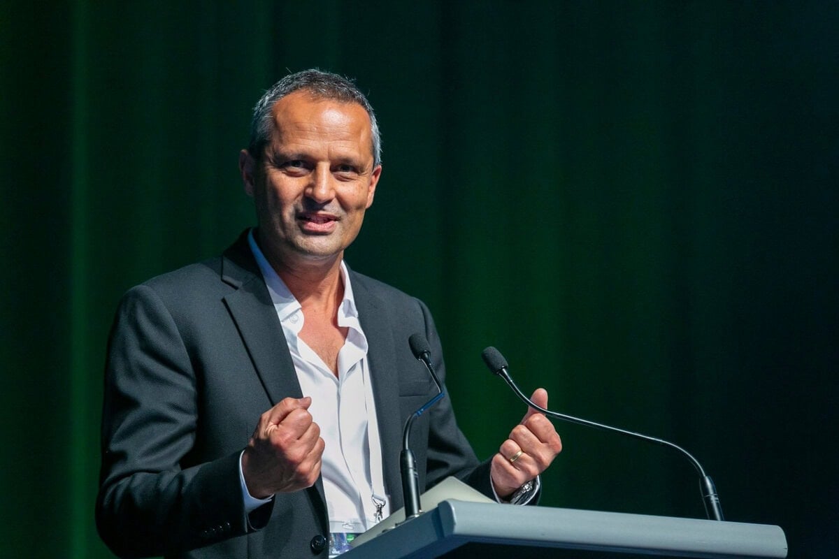 Хар костюмтай хүн хоёр микрофонтой индэр дээр ярьж байна. Тэр хоёр гараараа дохих бөгөөд дэвсгэр нь хар ногоон өнгөтэй. - Business.MN