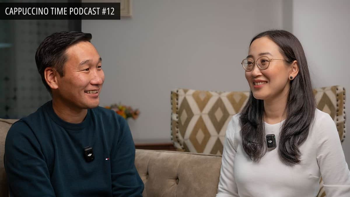 Эрэгтэй, эмэгтэй хүмүүс буйдан дээр сууж, микрофон зүүсэн байна. Тэд инээмсэглэж, харилцан яриа өрнүүлж, бүтээлч сэтгэлгээгээ харуулж байна. Текст дээр "Cappuccino Time Podcast EP12" гэж бичсэн байна. Төвийг сахисан өнгөт чимэглэл бүхий тав тухтай орчин. - Business.MN