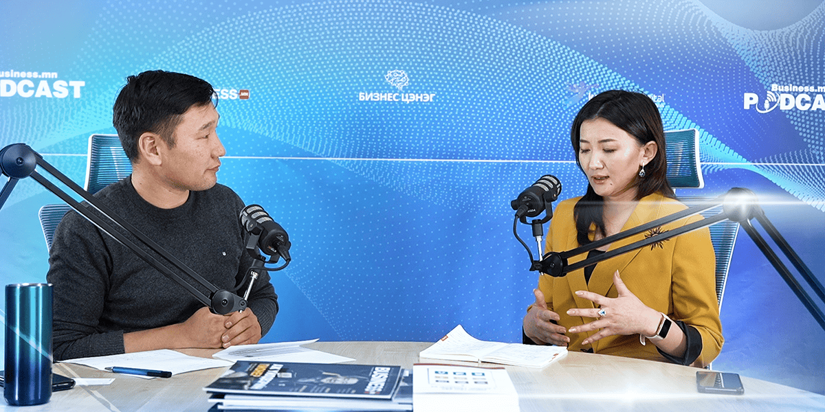 Хоёр хүн подкаст бичлэг хийх үед микрофоноор ярьж байна. Зүүн талд байгаа нэг эрэгтэй бичиг баримт барьдаг бол баруун талд байгаа эмэгтэй бизнес стратегийн талаар ярьж байхдаа дохио зангаа. Арын дэвсгэр нь цэнхэр өнгийн сэдэвтэй подкаст болон бизнесийн графикуудыг агуулна. Ширээн дээр ном гэх мэт янз бүрийн зүйл байдаг. - Business.MN