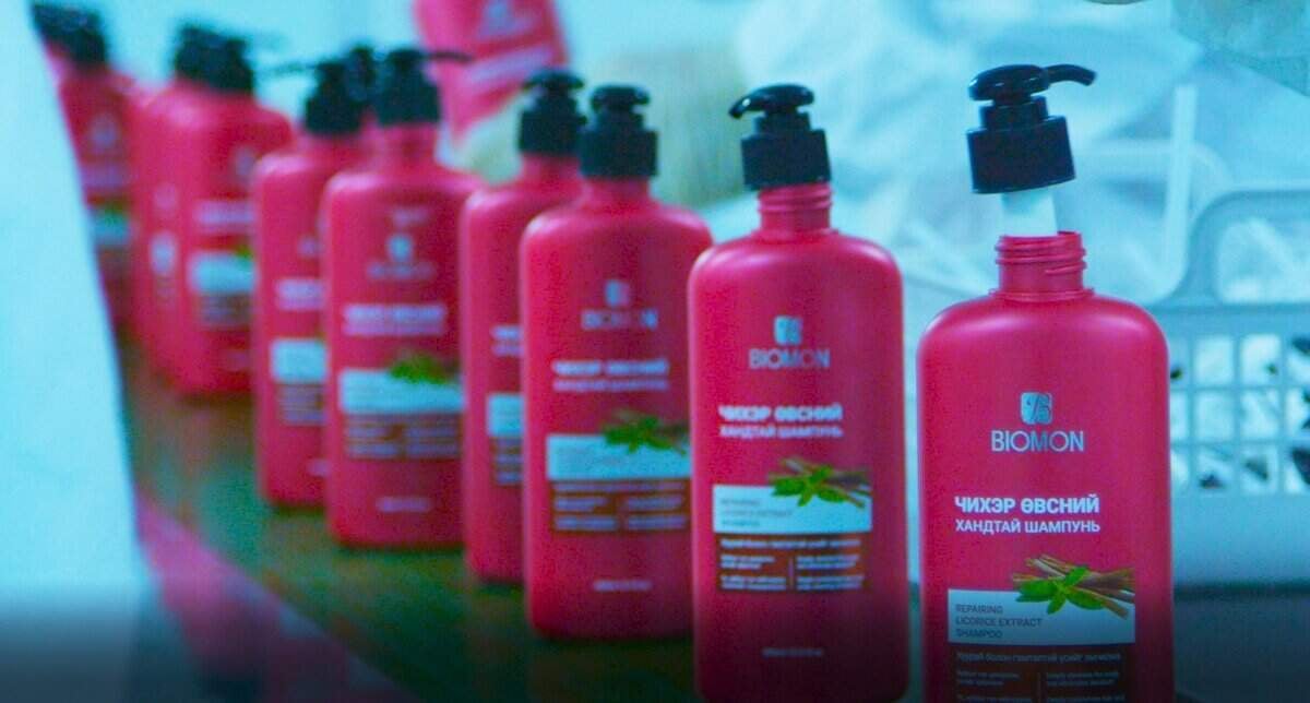 Тавиур дээр "Биомон" гэсэн шошготой хар шахуургатай улаан шампунийн лонх эгнүүлэн тавьжээ. Шошго нь ногоон навчны бичвэр, дүрсийг багтаасан бөгөөд энэ нь ургамлын гаралтай орцыг санал болгож байна. Арын дэвсгэр дээр бүдгэрсэн цагаан, цайвар цэнхэр өнгө нь аливаа бизнес эрхлэгчдэд тохиромжтой урам зоригтой уур амьсгалыг бүрдүүлдэг. - Business.MN