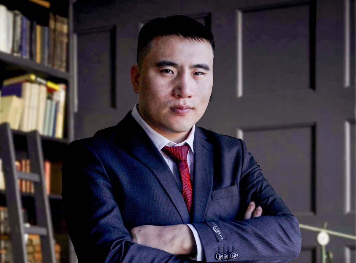 Костюм, улаан зангиа зүүсэн хүн бараан хавтантай хана, номын тавиурын өмнө гараа зөрүүлэн зогсож байна. Тохиргоо нь мэргэжлийн, албан ёсны харагдаж байна. - Business.MN