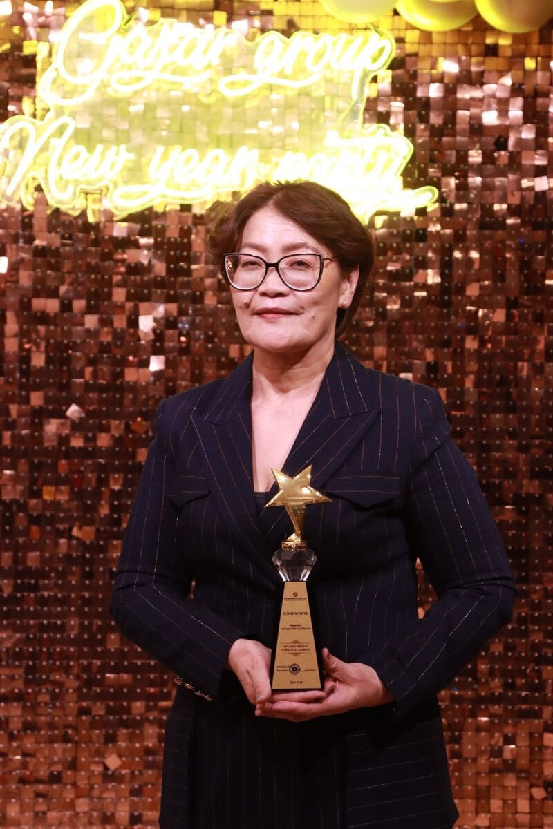 A person in a dark pinstripe suit holds a star-shaped trophy, standing in front of a shimmering backdrop with neon text.
