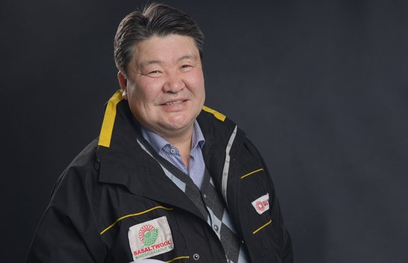 Богино хар үстэй, шар өнгийн өргөлт, лого бүхий хар хүрэм өмссөн бизнес эрхлэгч камер руу инээмсэглэж байна. Арын дэвсгэр нь энгийн бараан өнгөтэй бөгөөд тухайн хүнийг дүрсний гол анхаарал болгодог. - Business.MN