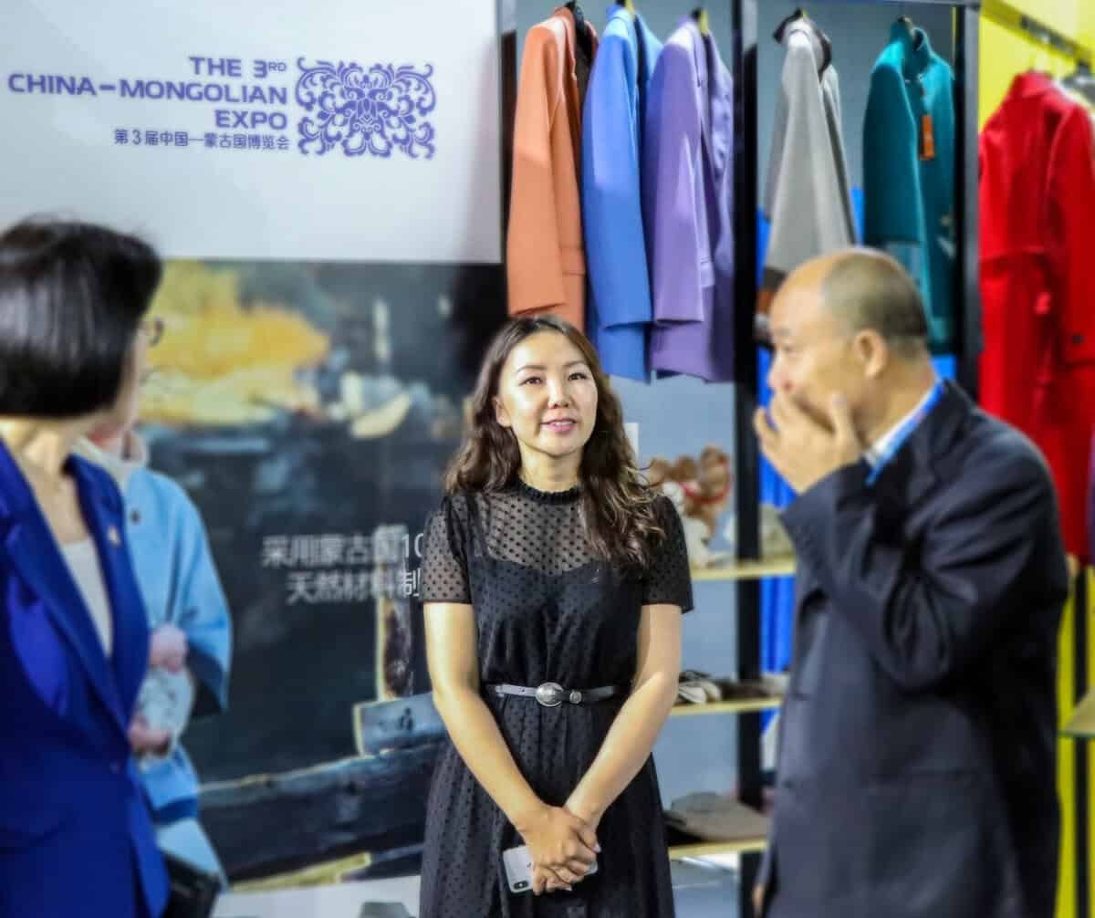 Хятад Монголын экспо дээр хар даашинз өмссөн эмэгтэй инээмсэглэн зогсож байна. Ойролцоох өөр хоёр хүн зогсож байгаагийн нэг нь цэнхэр хүрэмтэй, нөгөө нь бараан костюмтай. Ар талд нь өнгөлөг хувцас харагдана. - Business.MN