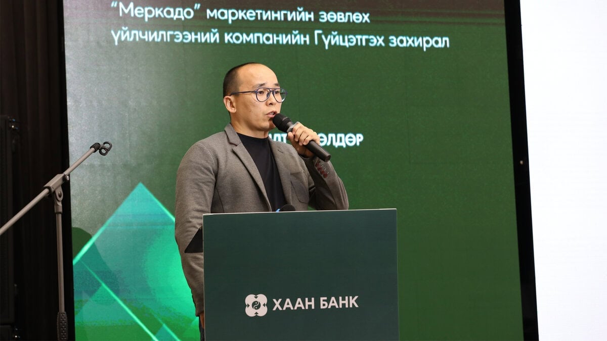 A man in glasses speaks into a microphone at a podium with the ХААН БАНК (Khan Bank) logo during an SME workshop: Бүтээгдэхүүн бус шийдэл санал болго, standing before a screen displaying text in Mongolian.