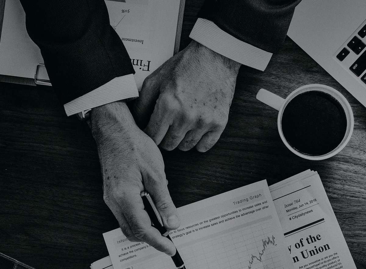 Костюм өмссөн энтрепренер ширээний ард суугаад санхүүгийн бичиг баримт, графикийг үзэж байна. Кофены аяга, зөөврийн компьютер болон бусад бичиг баримтыг ойролцоо байрлуулна. Зураг нь хар цагаан өнгөтэй бөгөөд тухайн хүн үзэг ашиглан аль нэг баримт бичгийг зааж байна. - Business.MN