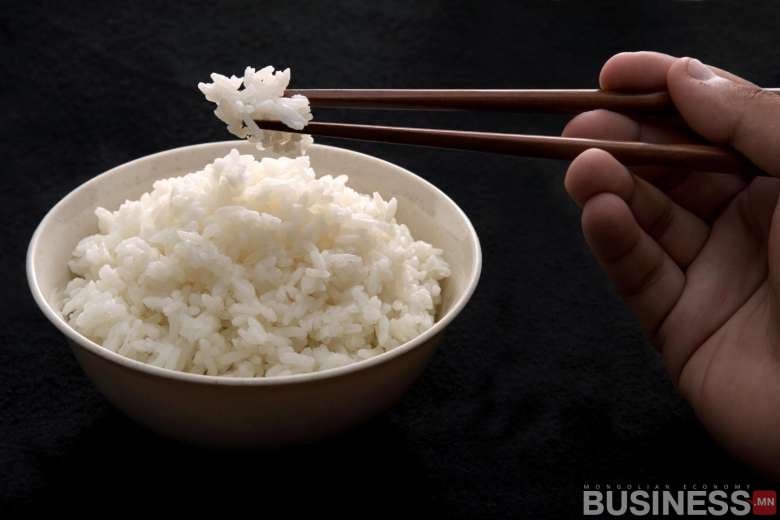 Уурын цагаан будаатай аяга нь харанхуй дэвсгэр дээр байрладаг. Нэг гар савх ашиглан аяганаас будаа өргөж байна. - Business.MN