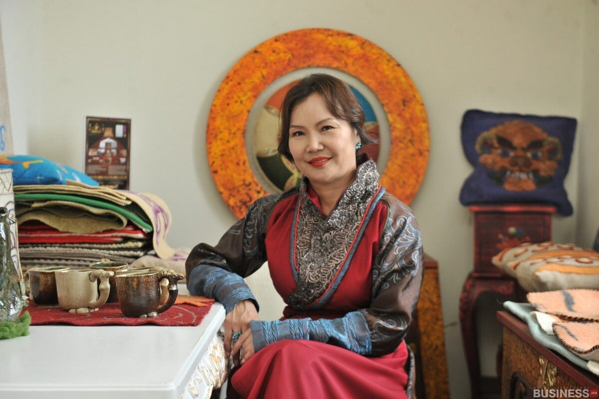 Нэхмэл эдлэл, дугуй тольоор чимэглэсэн өрөөнд вааран аягатай ширээний ард улаан, хар хувцастай эмэгтэй сууж байна. Хажууд нь овоолсон атираат даавуу, хатгамал дэрнүүдтэй камер руу инээмсэглэв. - Business.MN