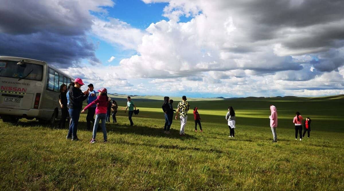 Багавтар үүлтэй тэнгэрийн дор уудам ногоон талбайд хэсэг хүмүүс зогсож байна. Зүүн талд нь "Ниссан" гэсэн бичигтэй цагаан өнгийн фургон байдаг. Тэд зарим нь алхаж, зарим нь хөдөлгөөнгүй зогсож, магадгүй бизнесийн боломж эсвэл бизнес эрхлэх санаагаа ярилцаж байгаа нээлттэй газар нутгийг таашааж байгаа бололтой. - Business.MN