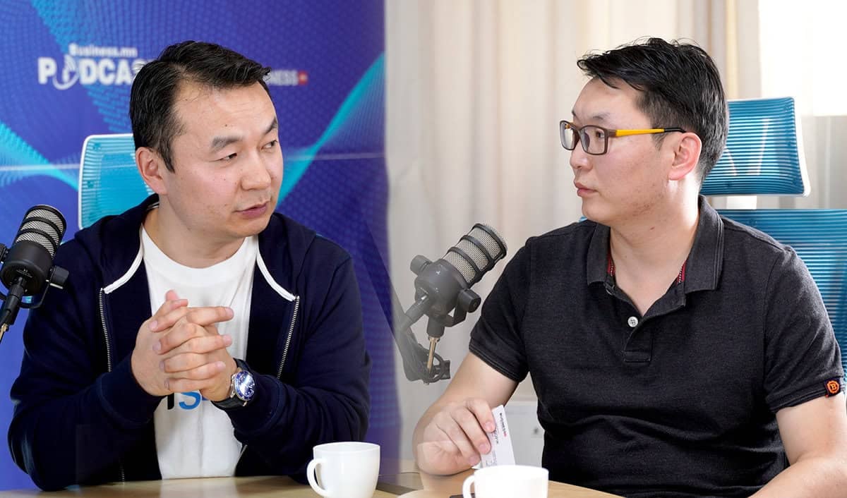 Хоёр эрэгтэй урд нь микрофонтой ширээний ард сууж байгаа нь подкаст бичиж буйг илтгэнэ. Зүүн талд байгаа хүн усан цэргийн хүрэмтэй, баруун талд байгаа хүн хар поло цамцтай. Ширээн дээр хоёр цагаан аяга байгаа бөгөөд цаана нь "бизнес" гэсэн подкаст лого харагдана. - Business.MN