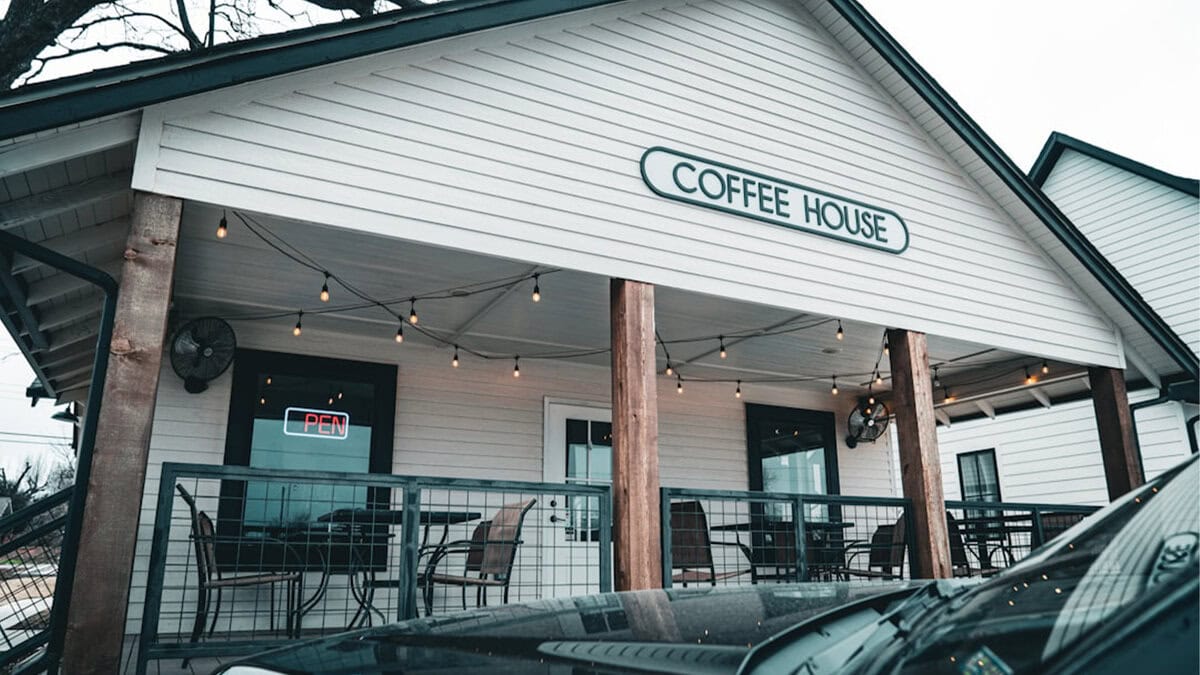 Орчин үеийн цагаан кофены байшин "Кофений байшин" гэсэн бичигтэй, утсан гэрэлтэй, гадаа суудалтай, цонхон дээрээ "НЭЭЛТТЭЙ" гэсэн бичигтэй. Урд талд нь машин зогсож байна. - Business.MN