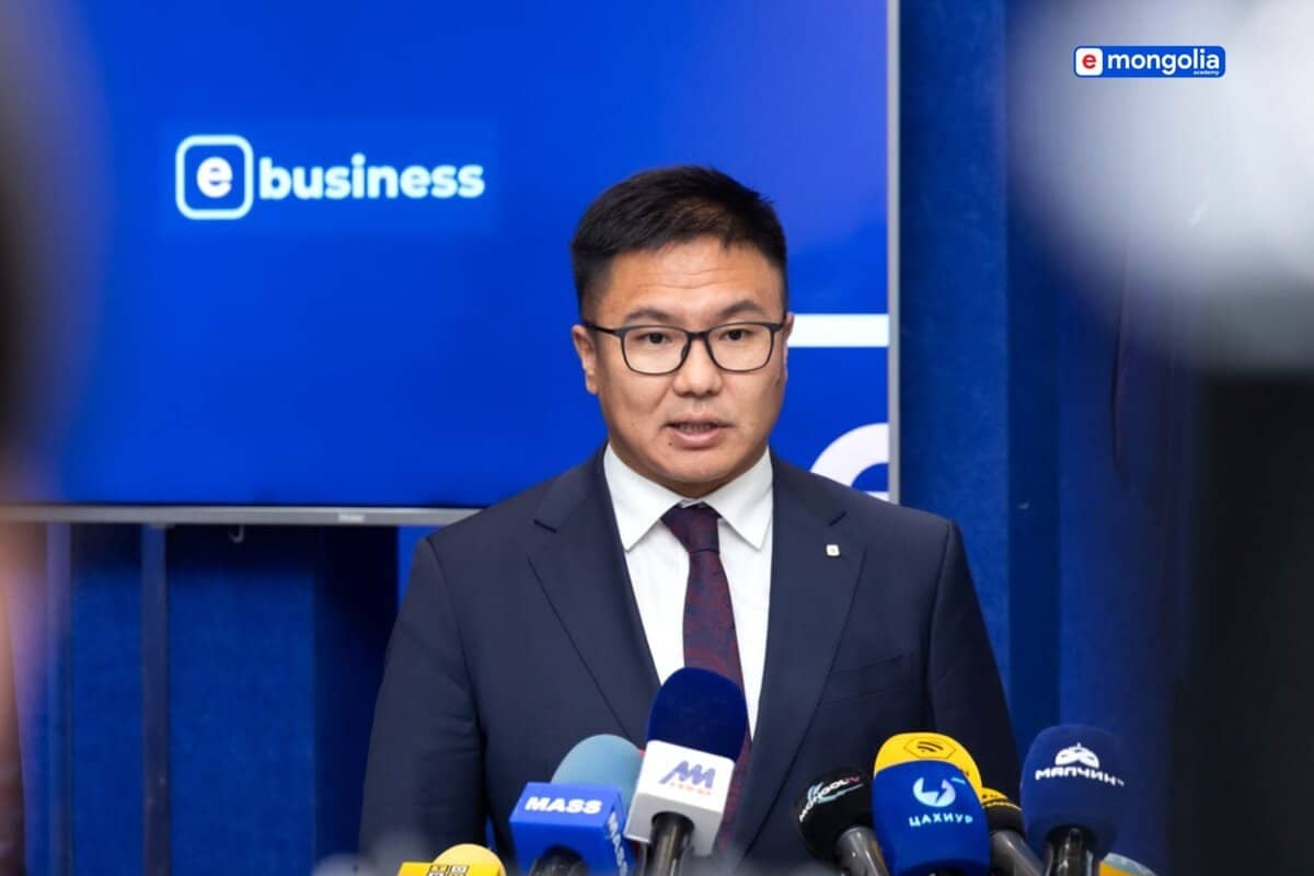 Костюм, зангиа зүүсэн эр урд нь олон микрофонтой индэр дээр ярьж байна. Цаана нь “цахим бизнес”, “цахим Монгол” гэсэн лого бүхий цэнхэр хана нь дижитал эдийн засагт нөлөө бүхий бизнес эрхлэгчийн үүргийг тодотгож байна. - Business.MN