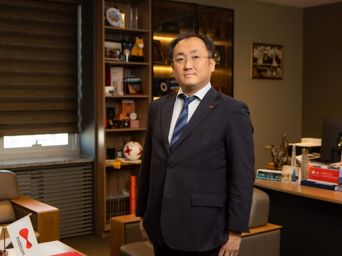 A man in a dark suit and tie stands in an office with shelves displaying awards and memorabilia, a window with blinds, and a desk with documents and a small flag in the foreground.