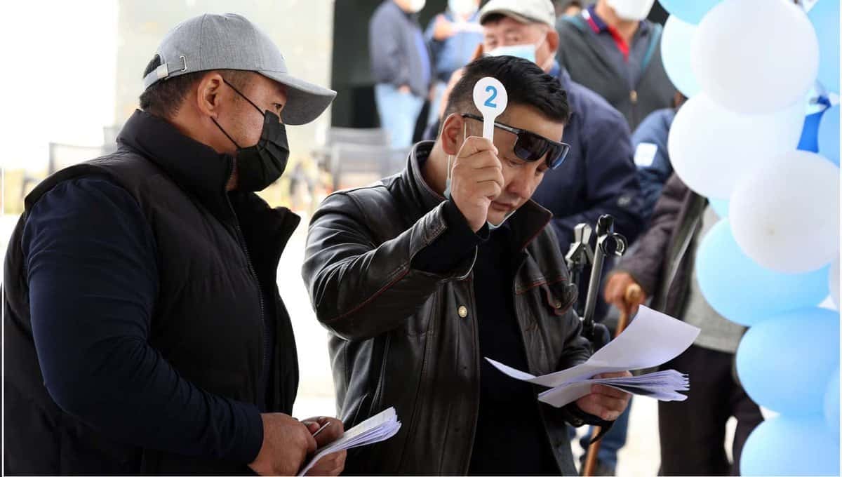 Эр хүн бусад хүмүүсээр хүрээлэгдсэн бичиг баримтыг шалгаж байхдаа '2' гэсэн тоотой тэмдэг барьж байна. Тэрээр хар савхин хүрэм, нарны шил, нүүрний маск өмсдөг. Урд талын нөгөө хүн хар малгай, нүүрний маск, бараан хүрэм өмсөж, цаас барьчихсан - хоёулаа анхаарлаа төвлөрүүлж байгаа мэт харагдаж байна. - Business.MN