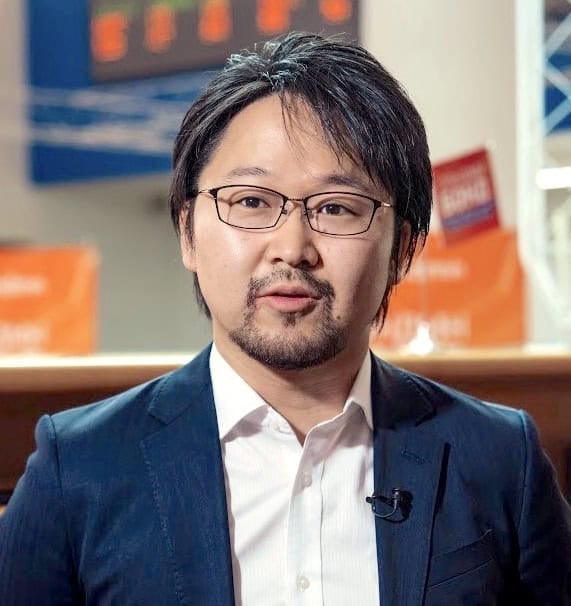 Костюм өмссөн нүдний шилтэй, нүүрний үстэй бизнес эрхлэгч улбар шар, цэнхэр өнгийн бүдгэрсэн дэвсгэртэй ярьж байна. - Business.MN