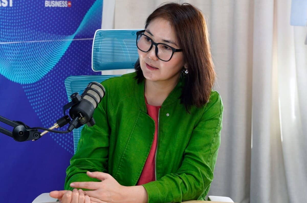 Дунд зэргийн урт хар үстэй, хар шилтэй хүн суугаад микрофоноор ярьж байна. Тэд ногоон хүрэм, улаан цамц өмссөн нь энтрепренерийн өөртөө итгэлтэй дүр төрхийг илэрхийлдэг. Ар талд нь хэсэгчлэн харагдах зарим текст бүхий цэнхэр туг байна. - Business.MN