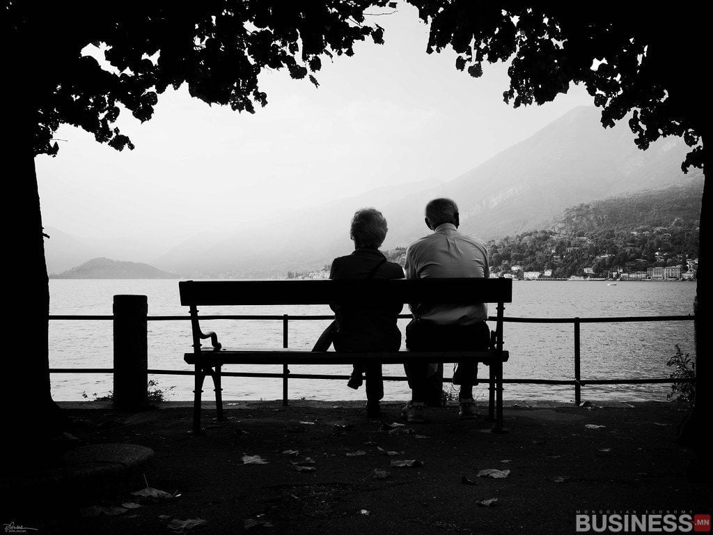 Ар талд нь уулстай нам гүм нуурыг ширтэж, сандал дээр сууж буй хоёр хүний хар цагаан зураг. Тэд модны сүүдэр дор тайван, тайван дүр төрхийг бий болгодог. - Business.MN