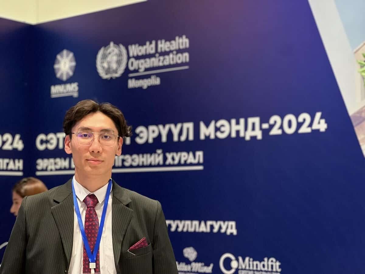 A man in a suit and tie stands in front of a blue backdrop with logos and text, including the World Health Organization and Mongolia. He is wearing glasses and a conference badge.