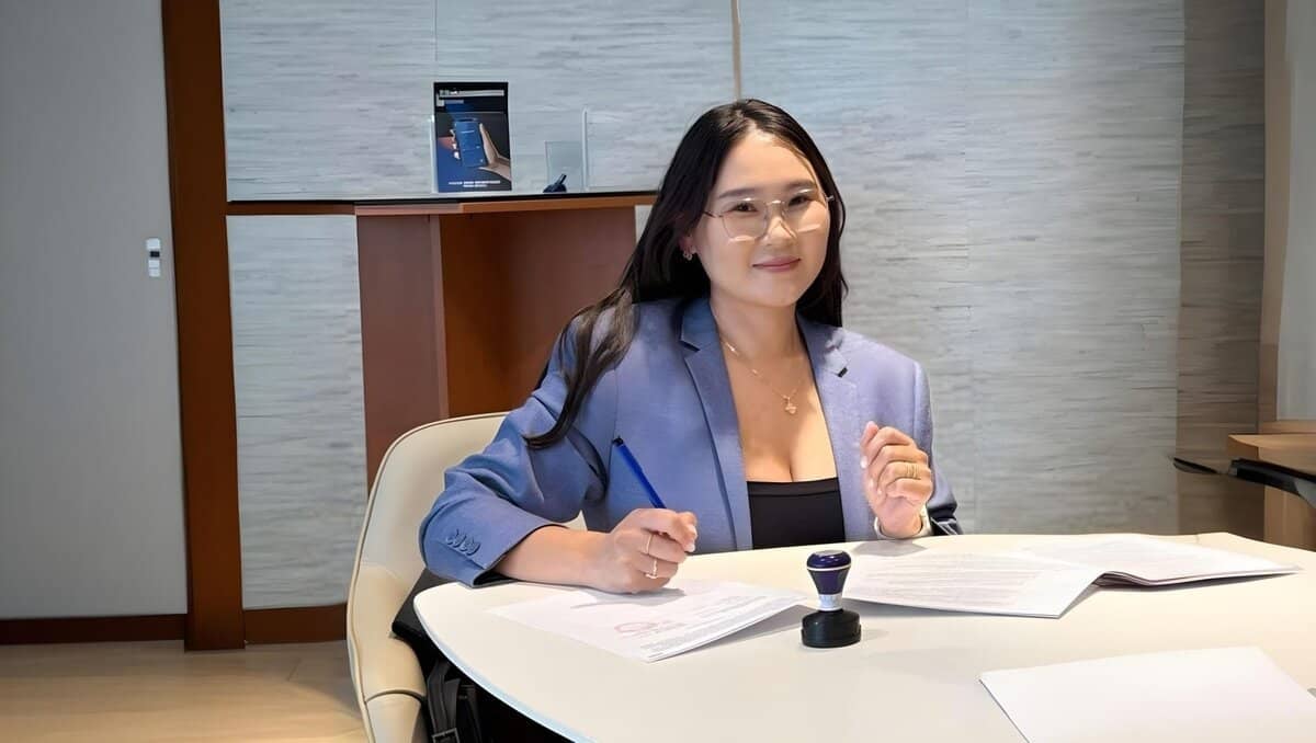 A woman with long black hair and glasses is seated at a desk, surrounded by paperwork and a stamp. Dressed in a light blue blazer, she smiles while holding a pen. Behind her, wooden shelves boast documents; she ambitiously plans to bring Тавилгын инновацыг Монголдоо угсарч олон улсад нийлүүлнэ to life.