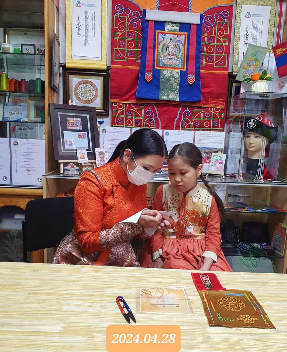 A woman in an orange traditional outfit and mask is seated at a table, assisting a young girl in a matching outfit with embroidery. They are surrounded by cultural artifacts and framed items. The date "2024.04.28" is displayed at the bottom.