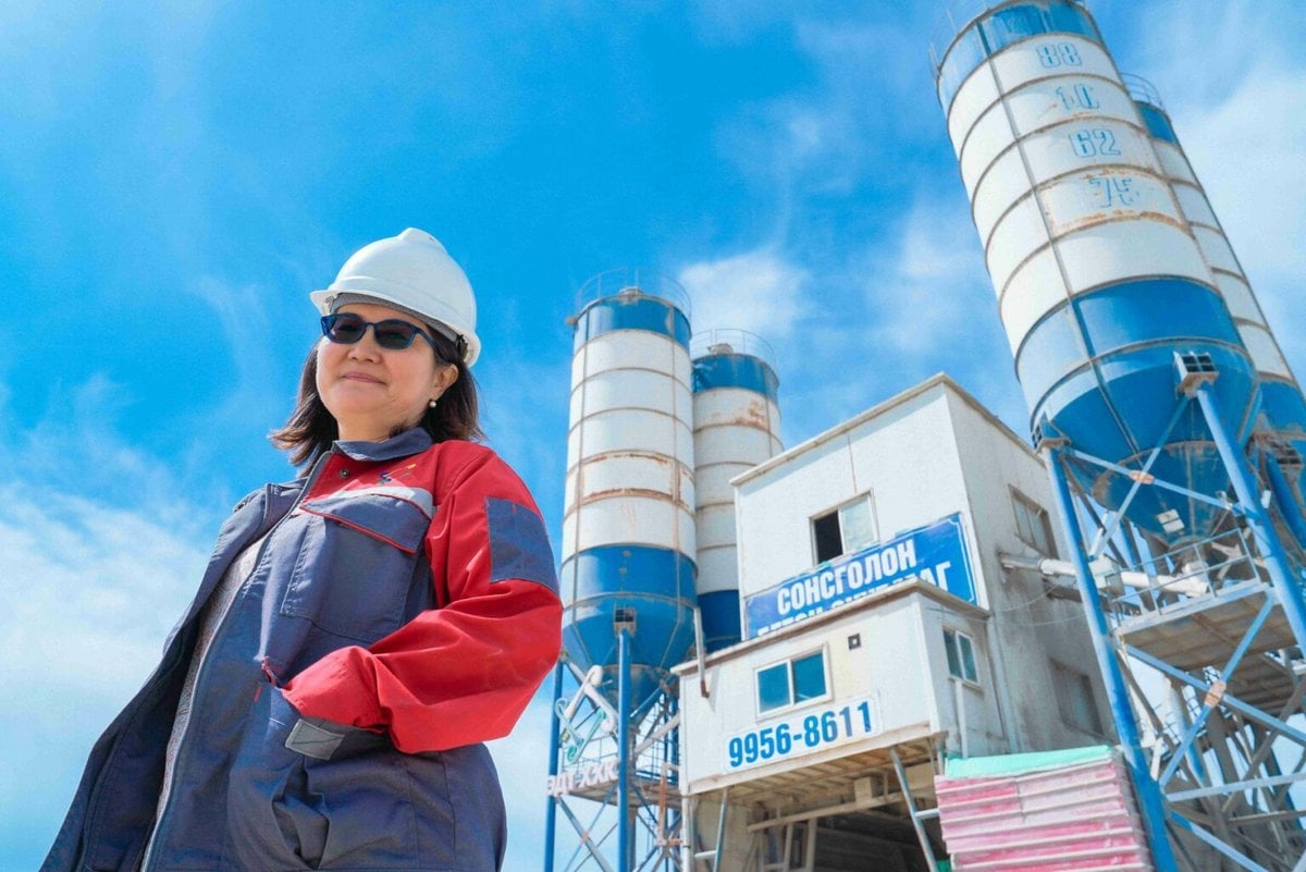 Цементийн үйлдвэрийн урд малгай, ажлын хүрэм өмссөн эмэгтэй ажилтны хөгжилд зориулсан бахархалыг илэрхийлэн, тод хөх тэнгэрийн дор сүндэрлэн боссон өндөр хөх цагаан силос бүхий итгэлтэй зогсож байна. - Business.MN