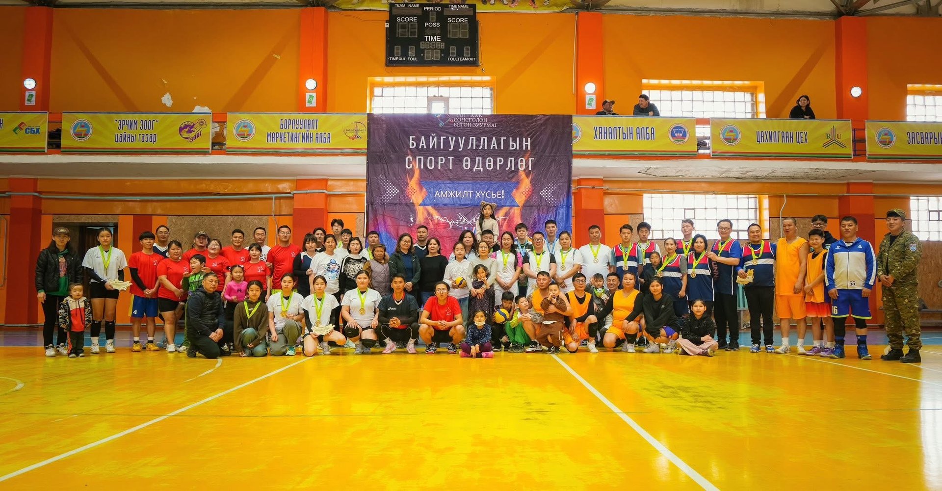 Спортын дүрэмт хувцастай, медальтай эрэгтэй, эмэгтэй хүмүүс тод гэрэлтүүлэгтэй битүү биеийн тамирын зааланд туг, самбарын өмнө хамтдаа зургаа авахуулж, спортын арга хэмжээг бахархалтайгаар тэмдэглэж байна. - Business.MN