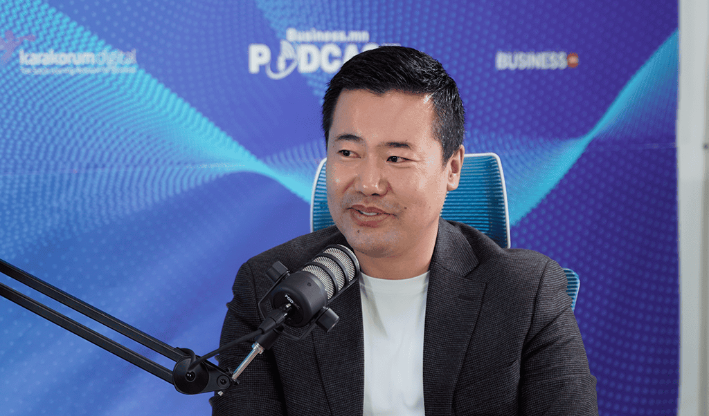 Харанхуй пиджак, цагаан цамц өмссөн эр микрофоны өмнө суугаад бизнесийн талаар догдолж ярьж байна. Арын дэвсгэр дээр "Business FM Podcast" гэсэн бичвэр бүхий цэнхэр туг болон холбогдох брэндийг харуулсан байна. - Business.MN