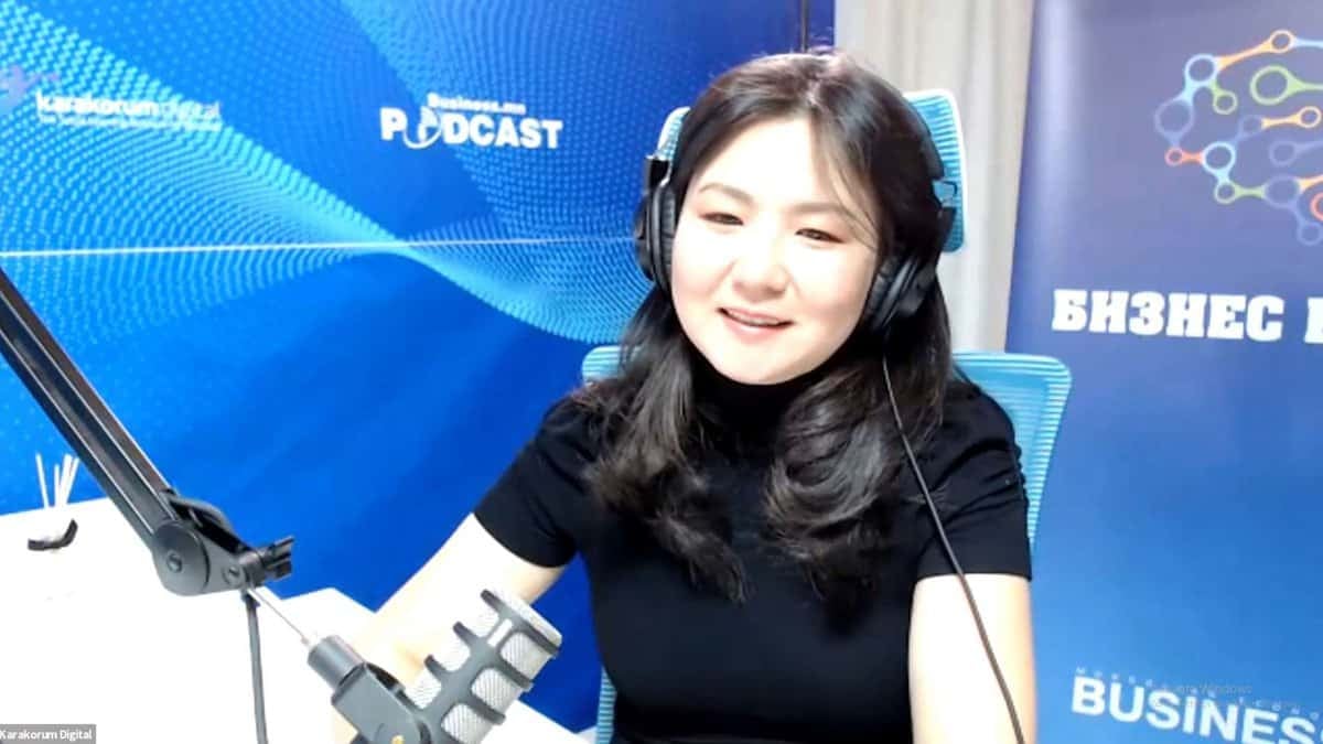 Чихэвч зүүсэн эмэгтэй микрофоны өмнө инээмсэглэн сууж байна. Тэр ар талд нь цэнхэр подкастын сэдэвтэй баннер бүхий студийн орчинд байна. - Business.MN