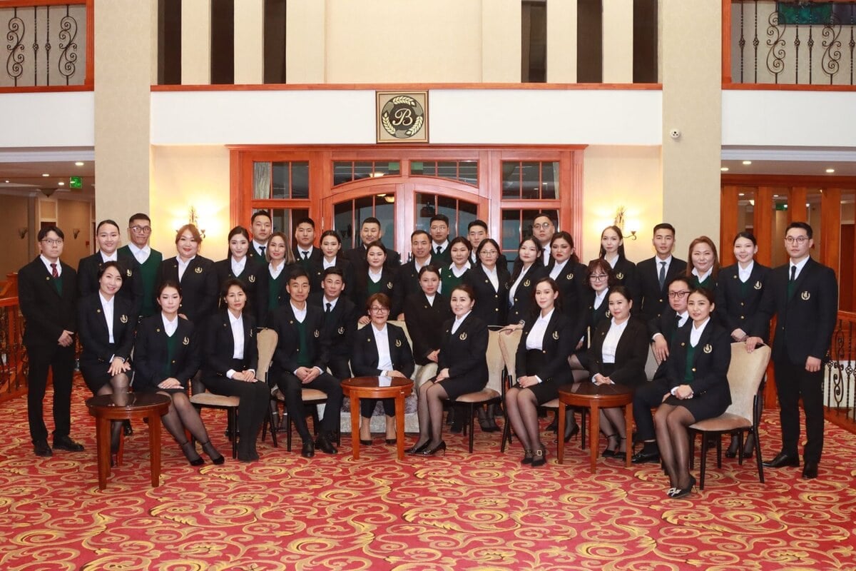 A group of 30 people dressed in formal black uniforms poses for a group photo in an elegant room with patterned carpet and a wooden backdrop. Most are standing, with a few sitting on chairs in front. The mood is professional and organized.