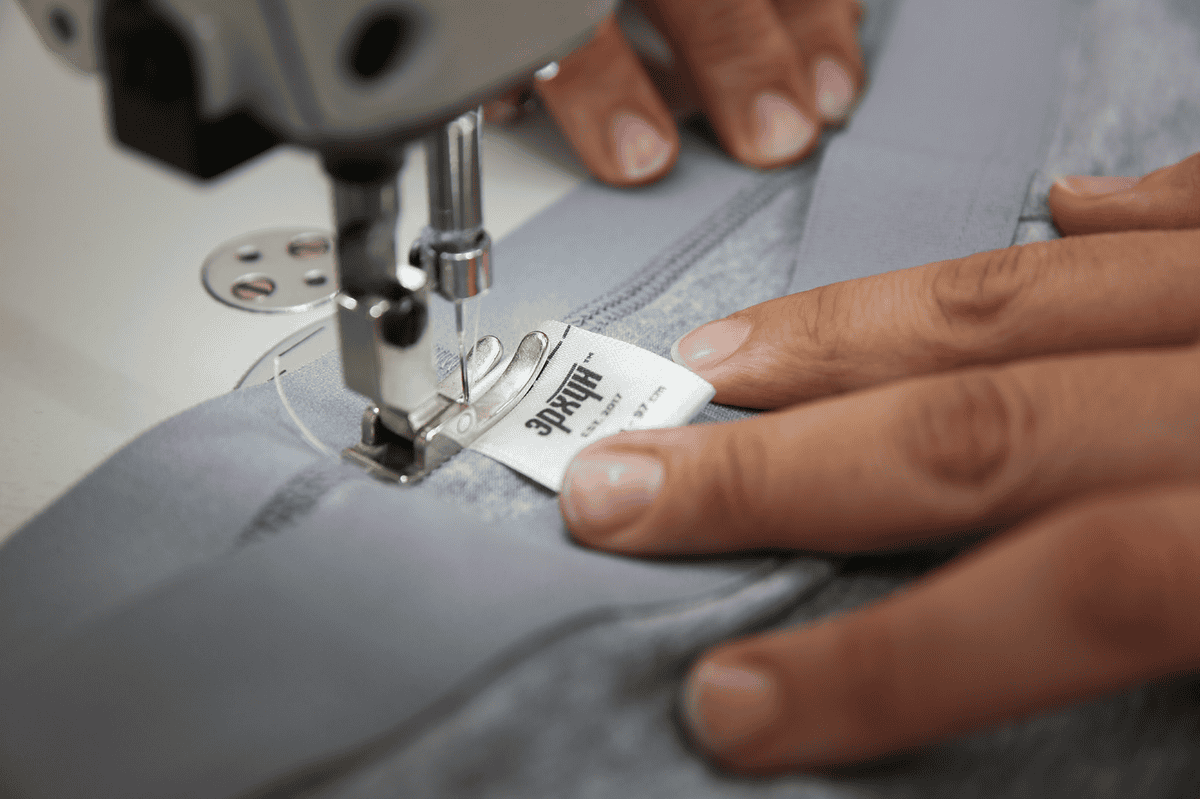 Оёдлын машин ашиглан саарал даавуун дээр шошго оёж буй гарууд нь аливаа компанид бизнес эрхлэхэд амжилтанд хүргэдэг нарийн гар урлалыг харуулж байна. - Business.MN