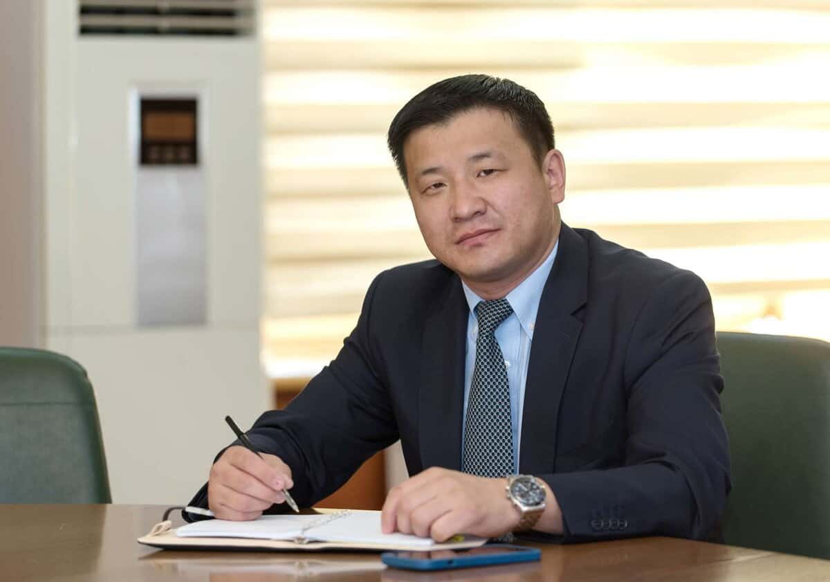 Харанхуй хувцастай хүн ширээний ард суугаад үзэг бариад тэмдэглэлийн дэвтэр дээр бичдэг. Тэмдэглэлийн дэвтрийн хажууд ширээн дээр утас байна. Арын дэвсгэр нь агааржуулагч, наалт бүхий цонхтой. Бизнес энтрепренер үл ялиг инээмсэглэн камер руу харж байна. - Business.MN