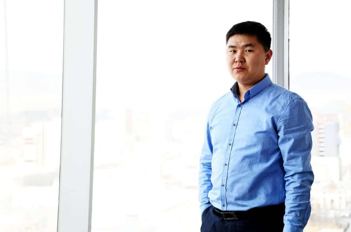 Цэнхэр цамц, бараан өмд өмссөн том цонхны дэргэд зогсож буй хүн. Гаднах хурц гэрэл нь тайван, мэргэжлийн уур амьсгалыг бүрдүүлдэг. Хотын өнгө үзэмж нь гэрэлд бүдэгхэн цонхоор үл ялиг харагдана. - Business.MN