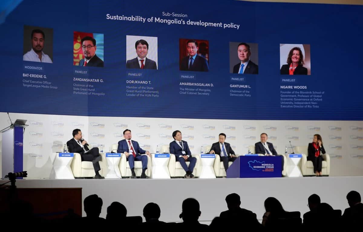 Албан ёсны хувцас өмссөн зургаан илтгэгчээс бүрдсэн самбар том дэлгэцийн өмнө тайзан дээр сууж, тэдний гэрэл зураг, гарчгийг харуулсан байна. Арын дэвсгэр дээр "Монгол Улсын хөгжлийн бодлогын тогтвортой байдал" гэж бичжээ. Үзэгчдийн гишүүд, тэр дундаа нэр хүндтэй энтрепренерүүд болон бизнесийн удирдагчид эхний эгнээнд сууж байна. - Business.MN