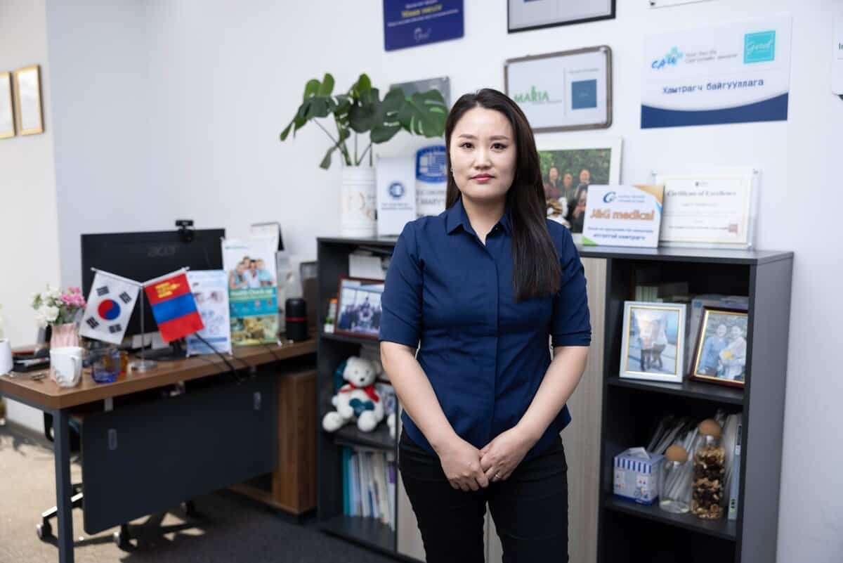 Цэнхэр цамцтай хүн амьдралын шинж тэмдгээр дүүрсэн оффист зогсож байна. Тэдний ард гэрэл зураг, гэрчилгээ, үнэ цэнийг хадгалсан ургамлуудыг байрлуулсан номын тавиур байдаг. Зүүн талд компьютер, туг далбаа, албан тасалгааны хэрэгсэл бүхий ширээ байрладаг бол хана нь жаазтай бичиг баримт, шагналуудыг харуулдаг. - Business.MN