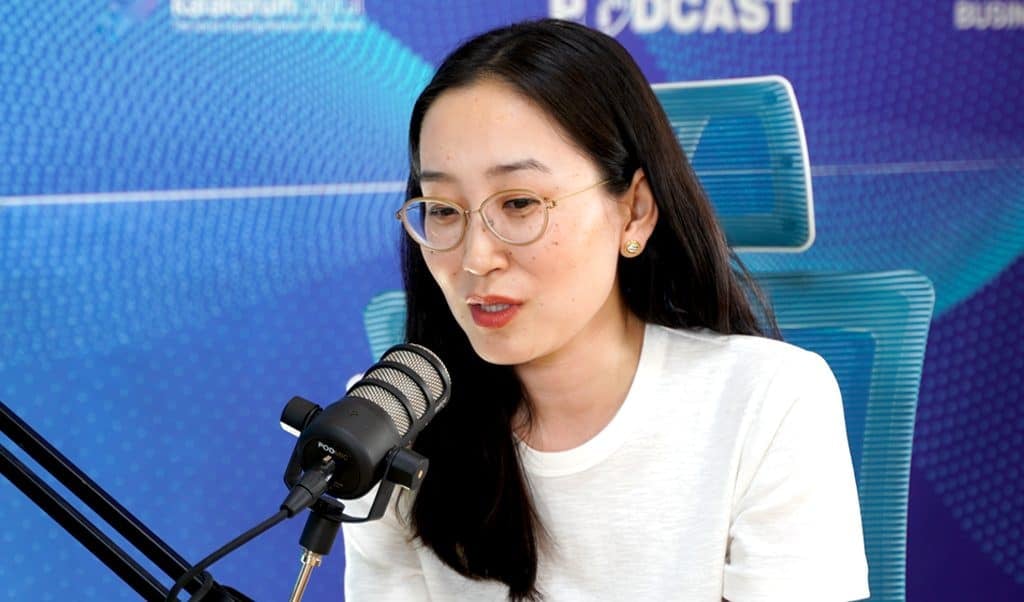 Урт хар үстэй, нүдний шилтэй эмэгтэй микрофоны өмнө суугаад ярьж байгаа бололтой. Тэрээр цагаан цамц өмсөж, цэнхэр дэвсгэр дээр ханан дээр хэсэгчлэн харагдахуйц бичвэртэй байх ба бизнесийн ертөнцөд энтрепренер болох аялалынхаа талаар ярилцаж байна. - Business.MN