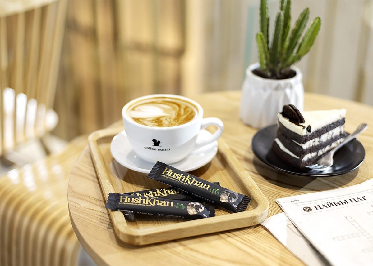 A wooden table with a cappuccino, a slice of layered cake on a black plate, three Hush Khan chocolate bars, a newspaper, and a small cactus in a white pot.