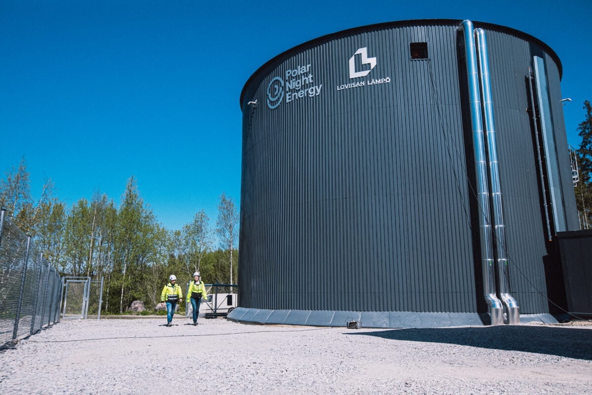 Two people in safety gear walk near a large cylindrical industrial building labeled “Polar Night Energy” and “Lounaisn Lampo,” surrounded by trees and a clear blue sky.
