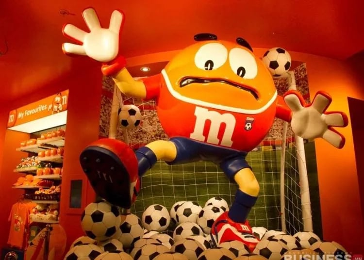 Хөлбөмбөгийн сэдэвтэй хувцас өмссөн улбар шар өнгийн том M&M дүр хөл бөмбөгөөр дүүрсэн өрөөнд зогсож, шинэ бизнесээ дөнгөж нээсэн мэт гайхширч байна. - Business.MN