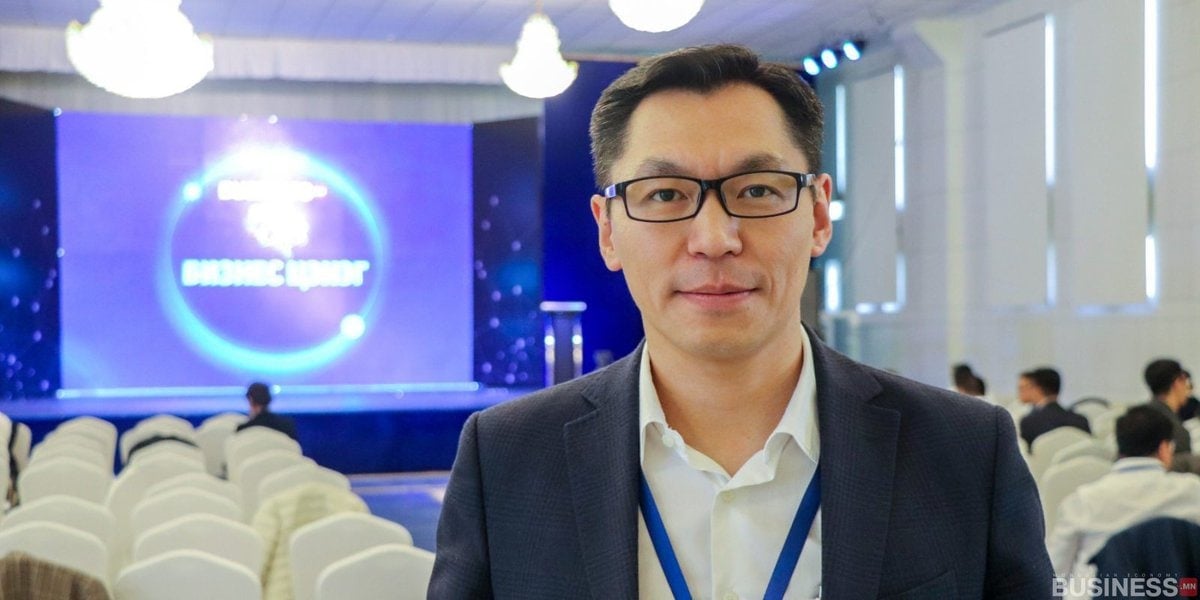 Костюм, нүдний шил зүүсэн хүн хурлын танхимд зогсож байна. Арын дэвсгэр дээр цэнхэр дэлгэцтэй тайз, цагаан сандал дээр сууж буй үзэгчид харагдаж байна. - Business.MN