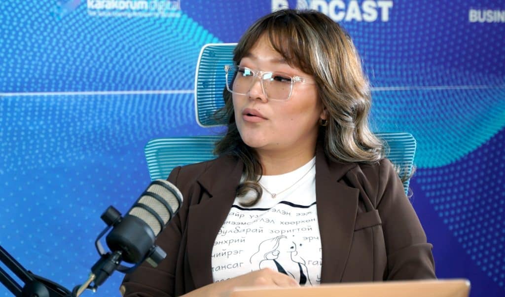 Мөрөн хүрсэн үстэй, нүдний шилтэй хүн микрофонд ярьж байна. Тэдгээр нь хийсвэр хээтэй, хэсэгчлэн харагдахуйц "PODCAST" гэсэн үг бүхий цэнхэр дэвсгэрийн өмнө суулгасан байна. Цагаан подволк, бор пиджак өмссөн энэ энтрепренер бизнесийн стратегийн талаарх ойлголтоо хуваалцдаг. - Business.MN