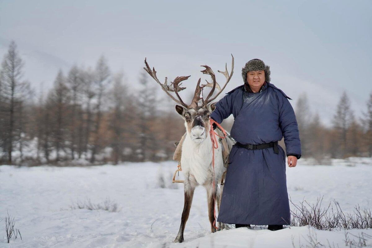 A person in a thick blue coat and fur hat stands beside a reindeer in a snowy landscape with bare trees in the background.