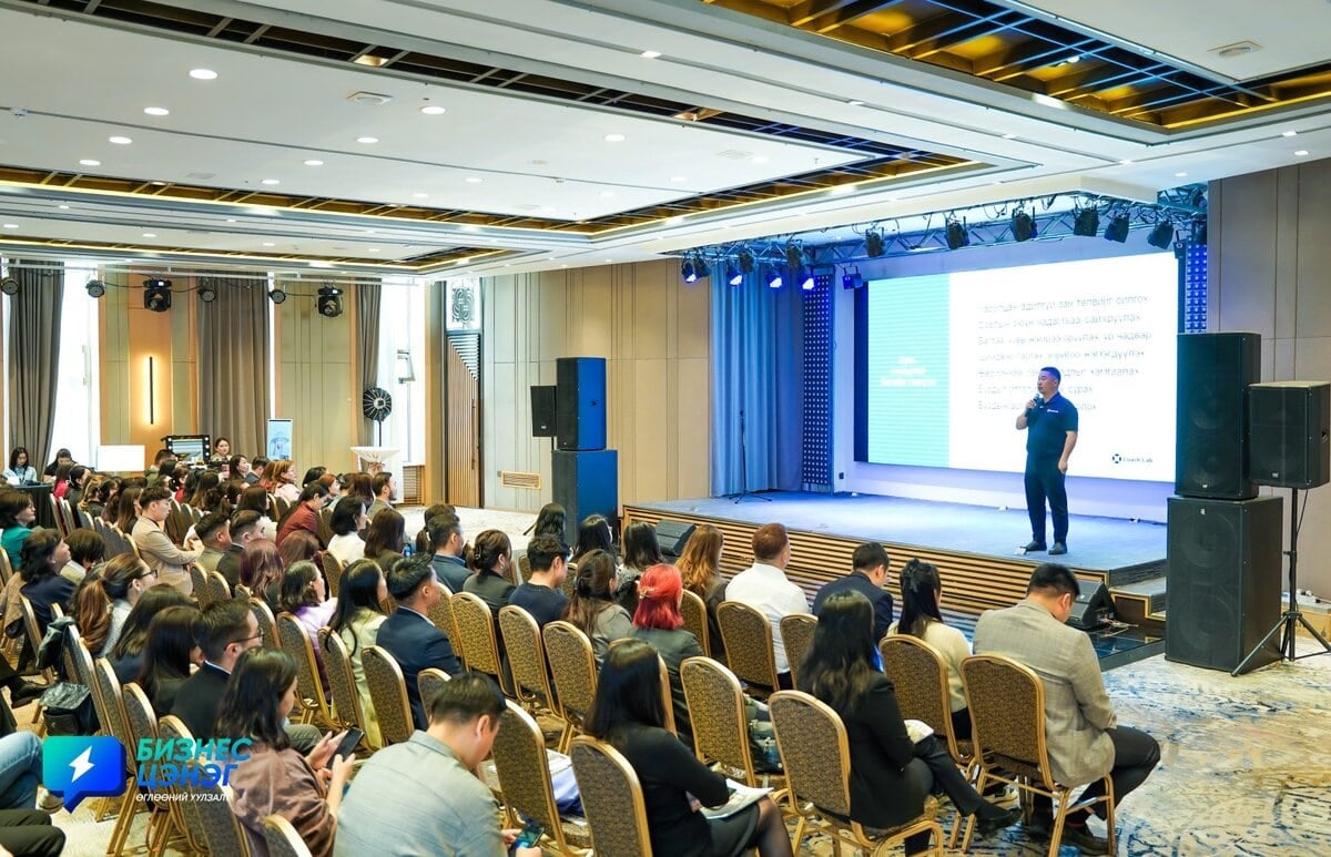 A speaker stands on stage presenting to a seated audience in a modern conference room. The presentation slide is projected on a large screen, and attendees are attentively listening.